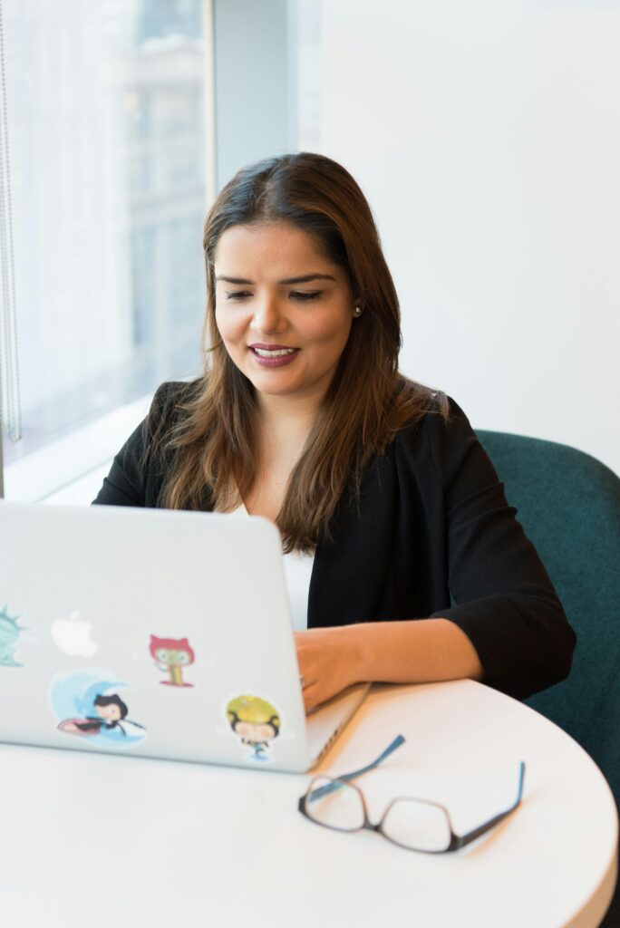 Smiling woman in black blazer working on laptop with open notebook on desk in a modern office setting.